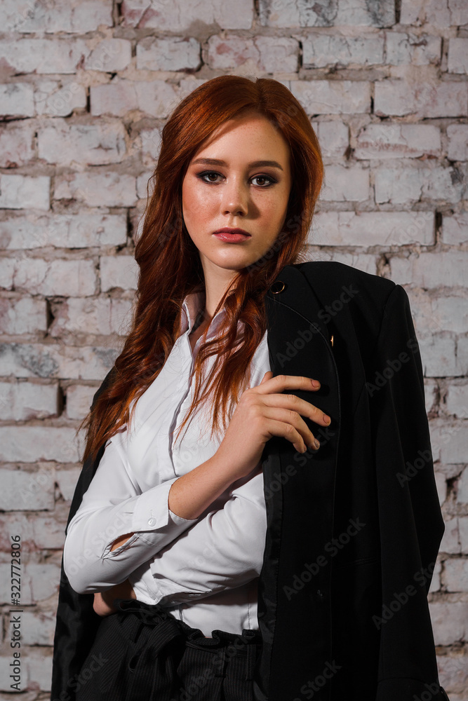 Young red-haired lady in a black suit consisted of trousers and jacket stands and looks directly. Self-confident business woman. White brick background.
