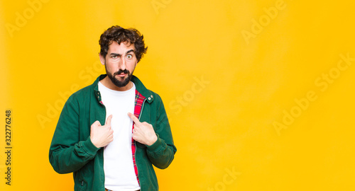 young crazy bearded man pointing to self with a confused and quizzical look, shocked and surprised to be chosen against flat wall photo