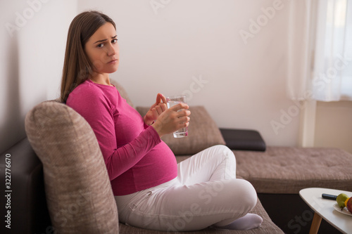 Sad pregnant woman drinking pill while sitting on sofa at her home.