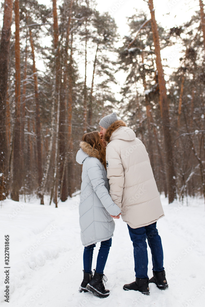 Fototapeta premium Happy lovers a guy and a girl who love each other hug, kiss, laugh, rage and walk in warm jackets in winter against the background of a snowy forest, a friendly family fun walk