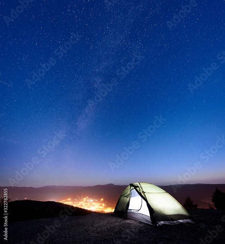 Tourist camping on the top of mountain in the morning. Illuminated tent under amazing night sky full of stars and Milky way. On the background beautiful starry sky, mountains and luminous town