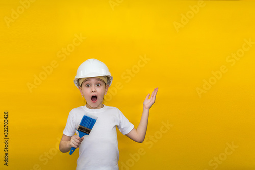 little girl with wide open eyes and mouth in a construction helmet holds a paint brush, points her finger at copyspace