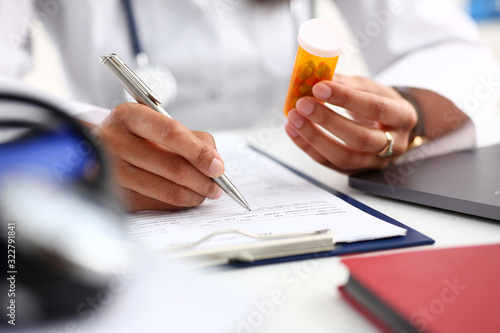 Fotografie Female black medicine doctor hand hold jar of pills and write prescription to patient at worktable