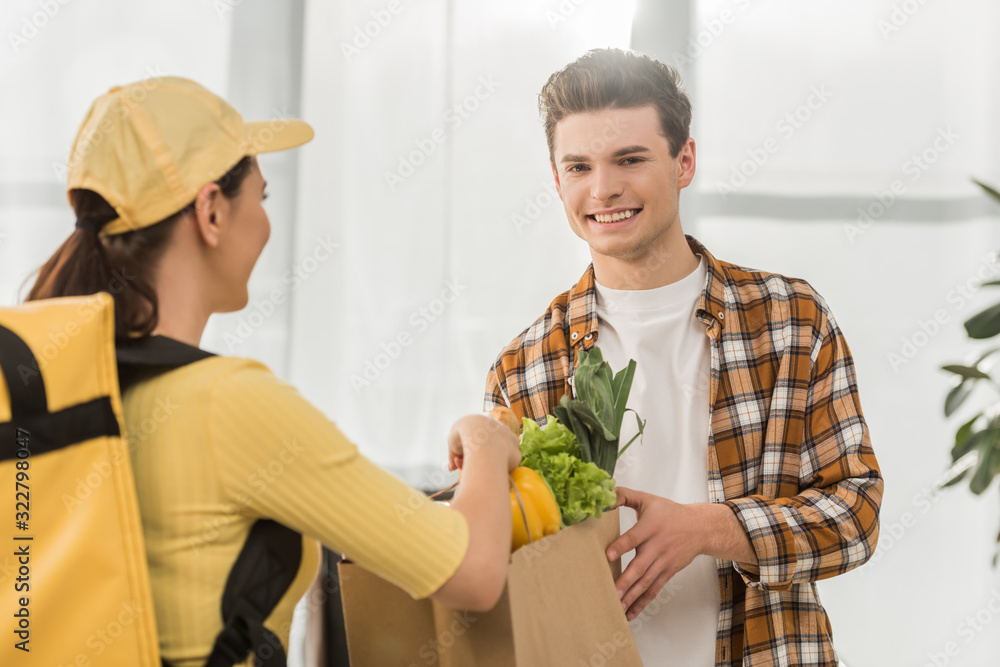 Selective focus of courier in yellow uniform giving package with fresh vegetables to smiling man Selective focus of courier in yellow uniform giving package with fresh vegetables to smiling man