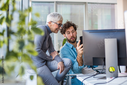 Two co-workers talking on a work break at the office. The man is showing something to the woman on his phone while she sits leaning against the window sill. photo