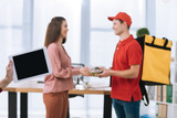 Selective focus of girl holding digital tablet while courier giving food in container to smiling businesswoman in office