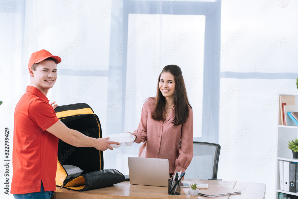 Delivery man with thermo bag giving food container to businesswoman and smiling at camera in office Delivery man with thermo bag giving food container to businesswoman and smiling at camera in office