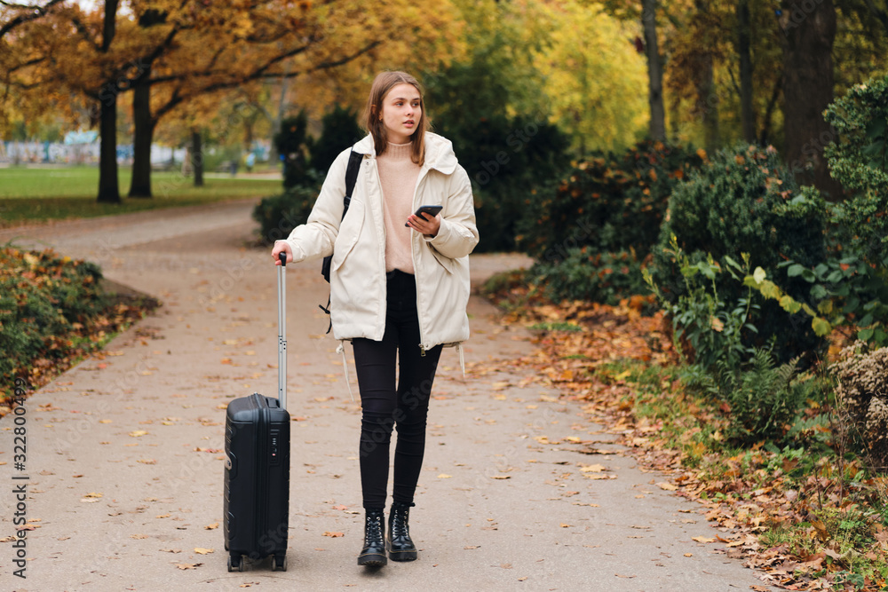 Fototapeta premium Attractive casual girl in down jacket with suitcase and cellphone walking through park