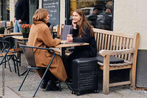 Two attractive girls happily talking during coffee break with suitcase in street cafe