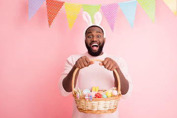 Photo of funny dark skin guy came easter party hold festive basket ginger bread painted eggs cookies wear sweater bunny ears colorful flags hang pastel pink color background