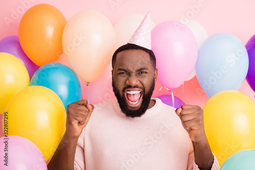 Photo of crazy funky dark skin guy enjoy birthday surprise raise fists surrounded many air balloons rejoicing wear paper cone cap sweater on pink color background photo