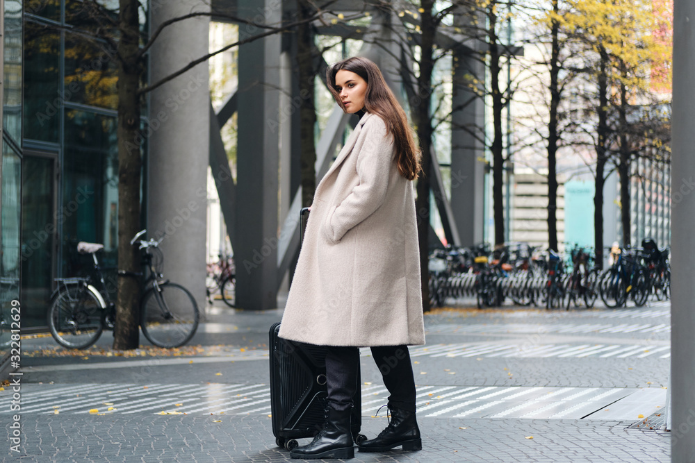 Attractive stylish girl in coat confidently looking aside standing on city street with suitcase