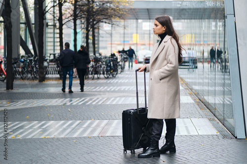 Side view of stylish girl in coat waiting on city street with suitcase