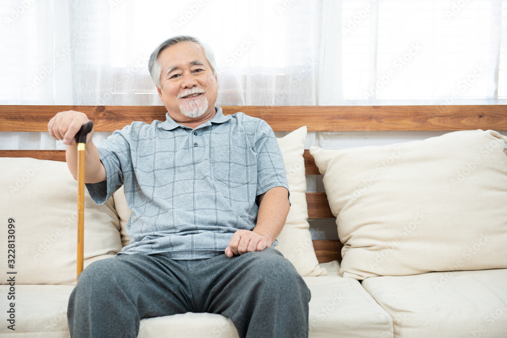 elderly old senior man sitting, resting his hands on wooden walking stick sitting on couch in living room in house after retirement.