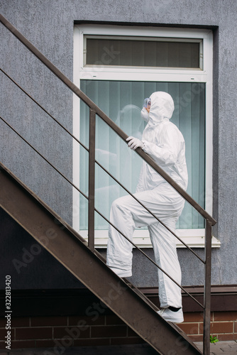 asian epidemiologist in hazmat suit and respirator mask walking upstairs outside building