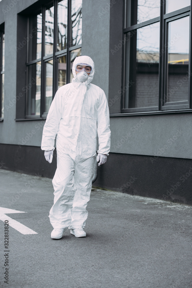 asian epidemiologist in hazmat suit and respirator mask looking at camera while standing on street near building
