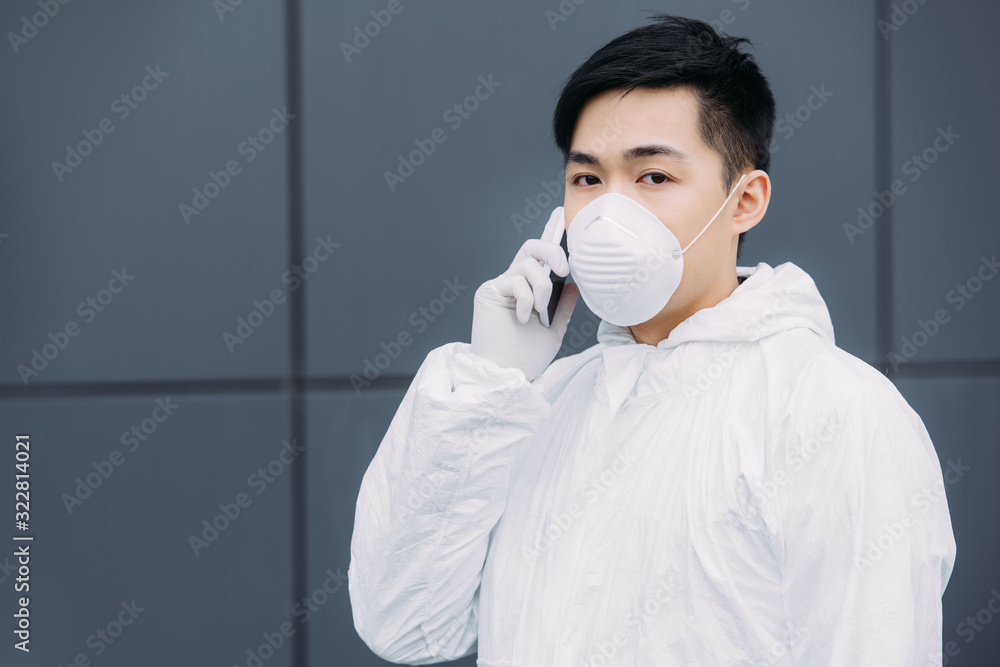 asian epidemiologist in hazmat suit and respirator mask talking on smartphone and looking at camera while standing on street asian epidemiologist in hazmat suit and respirator mask talking on smartphone and looking at camera while standing on street