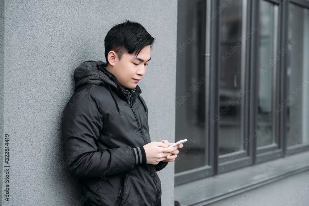 young asian man chatting on smartphone while standing by grey wall young asian man chatting on smartphone while standing by grey wall
