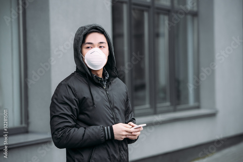 young asian man in respirator mask holding smartphone and looking away while standing on street