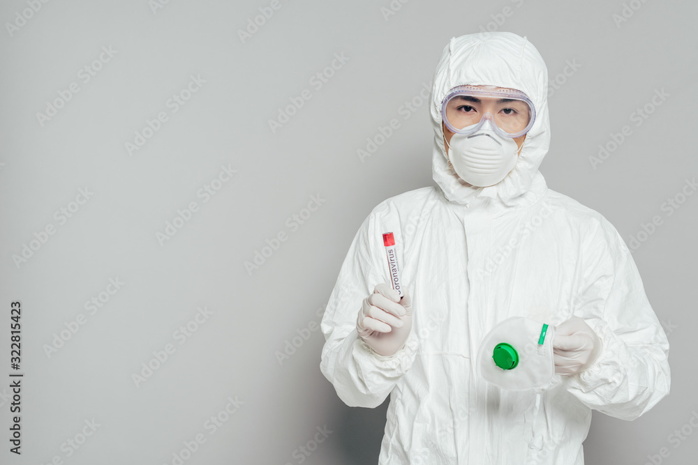 asian epidemiologist in hazmat suit looking at camera while holding respirator mask and test tube with blood sample on grey background asian epidemiologist in hazmat suit looking at camera while holding respirator mask and test tube with blood sample on grey background