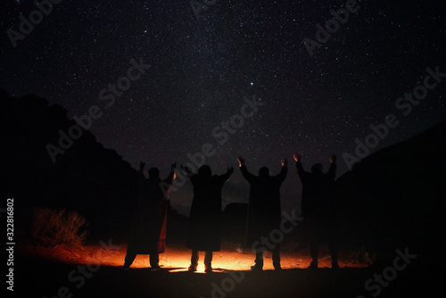 Four persons wearing long warm coats standing in night desert  hands raised to sky with stars  light on the ground  view from behind only silhouettes visible
