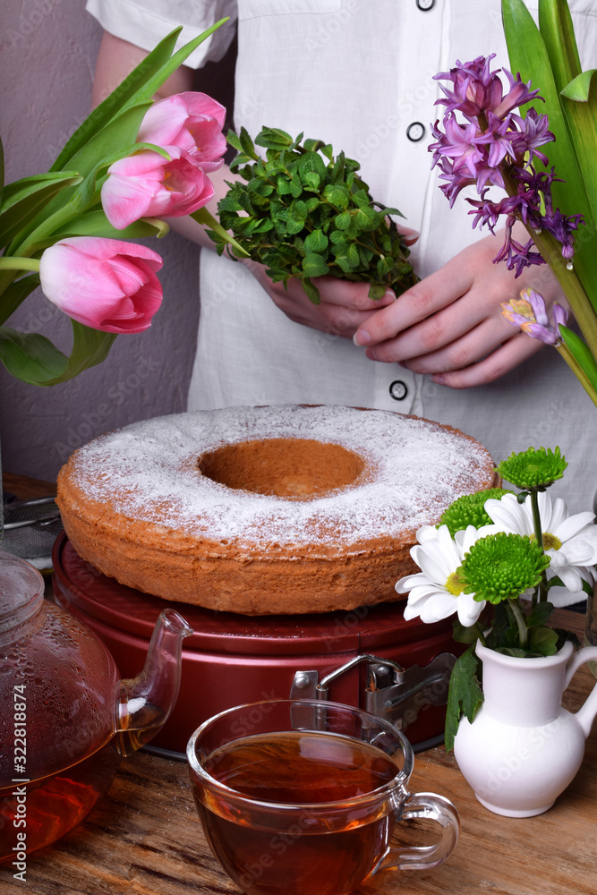 Sponge cake sprinkled with sugar powder surrounded by flowers. Woman wearing a white blouse in the background Sponge cake sprinkled with sugar powder surrounded by flowers. Woman wearing a white blouse in the background