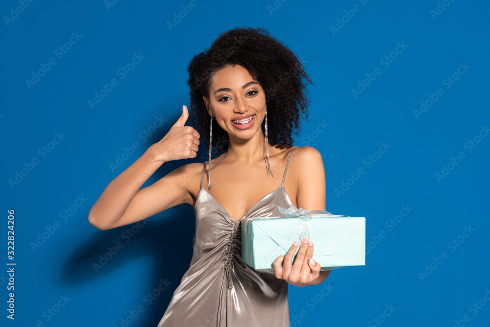 Obraz premium happy beautiful african american woman in silver dress holding gift and showing thumb up on blue background