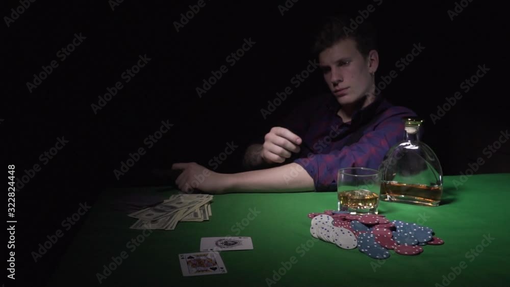 A young man with a cigar at a card table in a casino. A guy throws cards on a green table in a casino. On the table is a bottle of alcohol, a glass and playing chips. Moving camera. Slow motions A young man with a cigar at a card table in a casino. A guy throws cards on a green table in a casino. On the table is a bottle of alcohol, a glass and playing chips. Moving camera. Slow motions