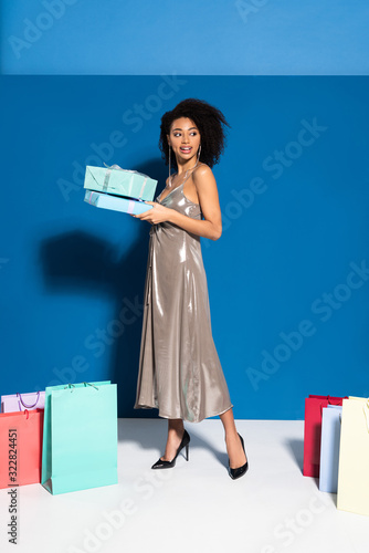 happy beautiful african american woman in silver dress holding gift boxes near shopping bags on blue background