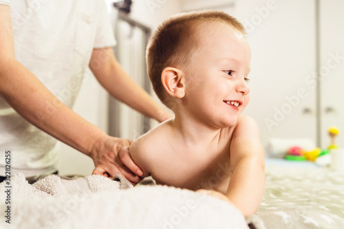 Small caucasian boy laying on the bed naked having back massage by professional female physiotherapist masseuse in a rehabilitation center salon or home office of physical therapy