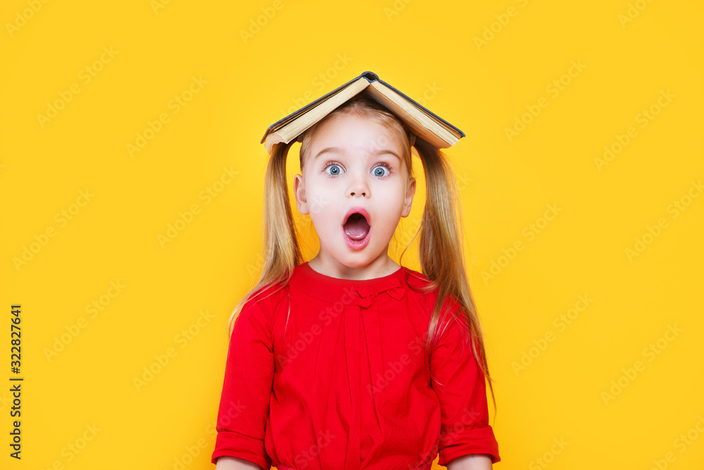 Shocked little girl holding book on her head and looking at camera - obrazy, fototapety, plakaty 