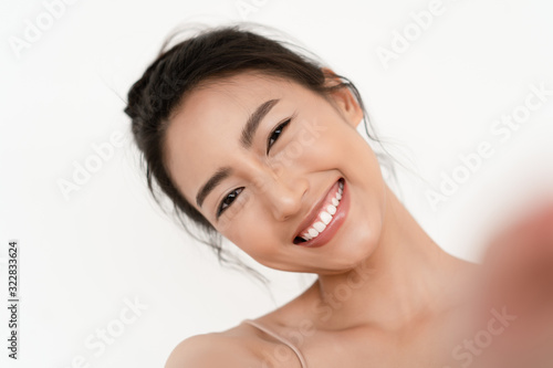 Closeup portrait of beautiful young Asian smilling with white teeth and taking selfie of herself over white wall background. Natural Beauty and Healthy Woman.