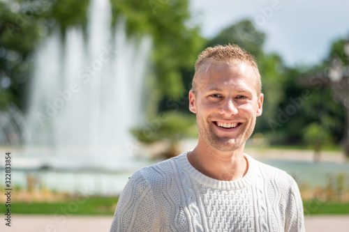 Portrait beau gosse charmant jeune homme caucasian blond aux yeux bleu joyeux et confiance en soi séduisant chanceux charismatique européen heureux zen à motivation pull blanc jardin de la fontaine