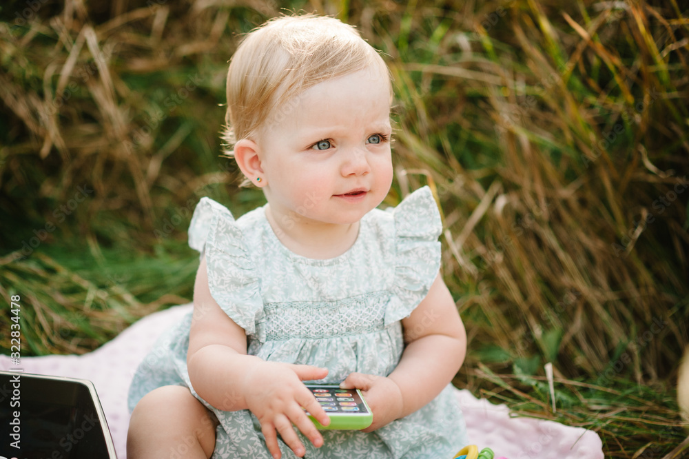 Cute baby girl sitting on blanket on the grass in the park . Summer portrait of beautiful daughter. Happy child playing toys in wild yellow field. Concept of family holiday, spring time together. Cute baby girl sitting on blanket on the grass in the park . Summer portrait of beautiful daughter. Happy child playing toys in wild yellow field. Concept of family holiday, spring time together.