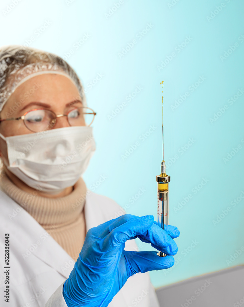 A small injection syringe in the doctor’s hand in a latex medical glove and a face mask. Virus protection concept. Medical theme on a light turquoise background. A small injection syringe in the doctor’s hand in a latex medical glove and a face mask. Virus protection concept. Medical theme on a light turquoise background.