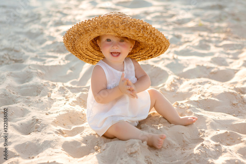 baby girl in white clothes and a straw hat sits on the white sand on the beach in summer © КРИСТИНА Игумнова