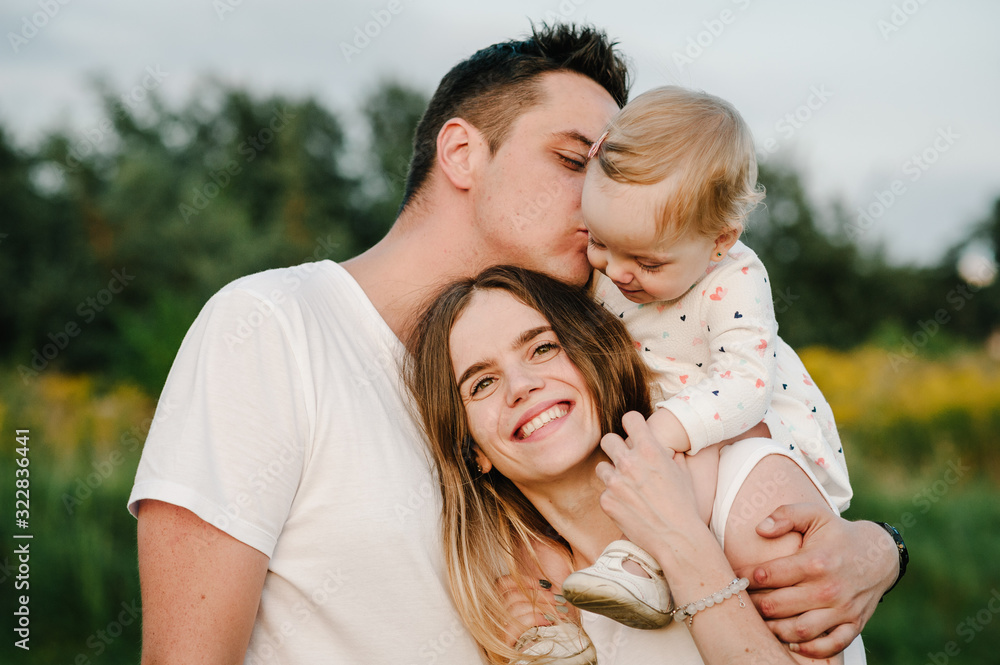 The daughter hugging parents on nature. Mom, dad and girl toddler, walk in the grass. Happy young family spending time together, outside, on vacation, outdoors. The concept of family holiday. The daughter hugging parents on nature. Mom, dad and girl toddler, walk in the grass. Happy young family spending time together, outside, on vacation, outdoors. The concept of family holiday.