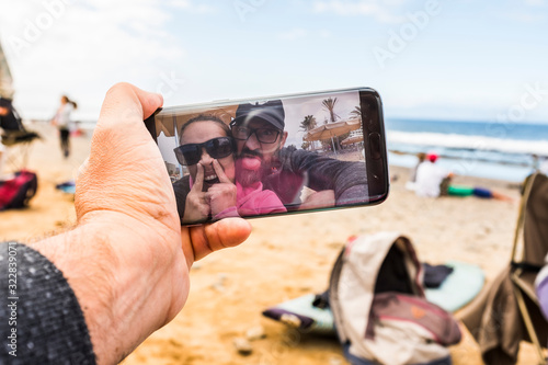 Man point of view of funny crazy couple having fun taking selfie or doing video conference call from the beach using a modern technology connected smart phone