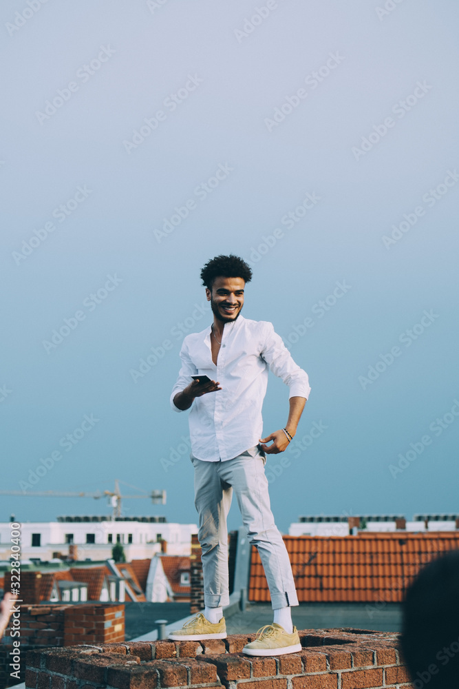 Smiling man using smart phone while standing on terrace against sky Smiling man using smart phone while standing on terrace against sky