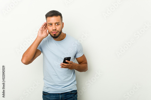 Young mixed race asian man holding a phone trying to listening a gossip. © Asier