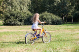 Young girl in white dress riding her bicycle in summer park on meadow with green grass