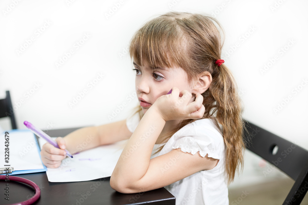 Little Caucasian girl with sad look sitting at the desk with pen in hand, boring lesson, white background