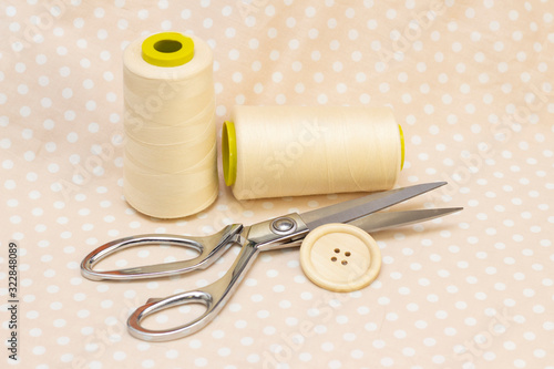 spools of beige cotton. scissors and a wooden button on a beige polka dot fabric. natural sewing concept