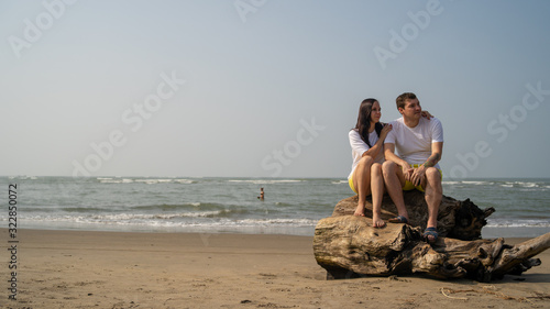Happy couple posing on driftwood near sea. Loving couple hugging during date on beach against waving sea and cloudless sky.