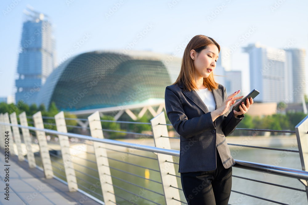 Fototapeta premium Young Asian business woman with Phone standing outdoors using smartphone in modern city financial district. Business and Communication concept.