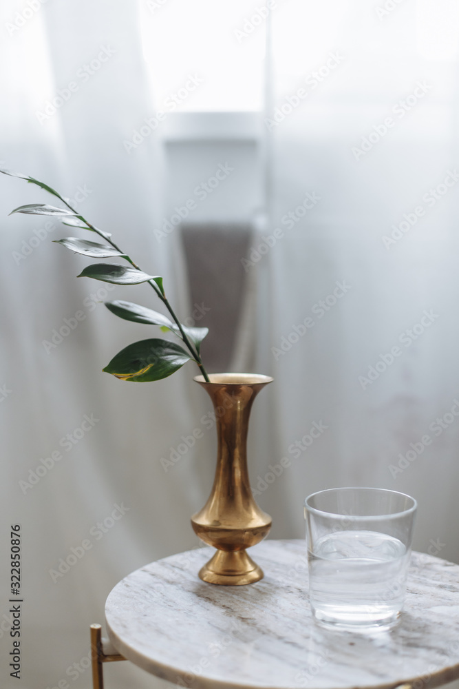 Gold vase with garnish with greens marble backsplash wooden board white drapes glass water in cozy scandinavian interior Gold vase with garnish with greens marble backsplash wooden board white drapes glass water in cozy scandinavian interior