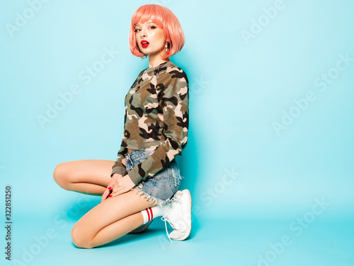 Portrait of young beautiful hipster bad girl in trendy red summer clothes and earring in her nose.Sexy carefree smiling woman sitting in studio in pink wig near blue wall.Positive model having fun