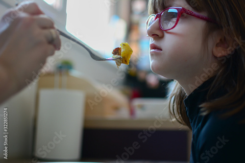 Close-up, side view of Cute little girl dislikes healthy lunch with her mother. Adorable girl looking seriously at the meat on her fork. Right nutrition.