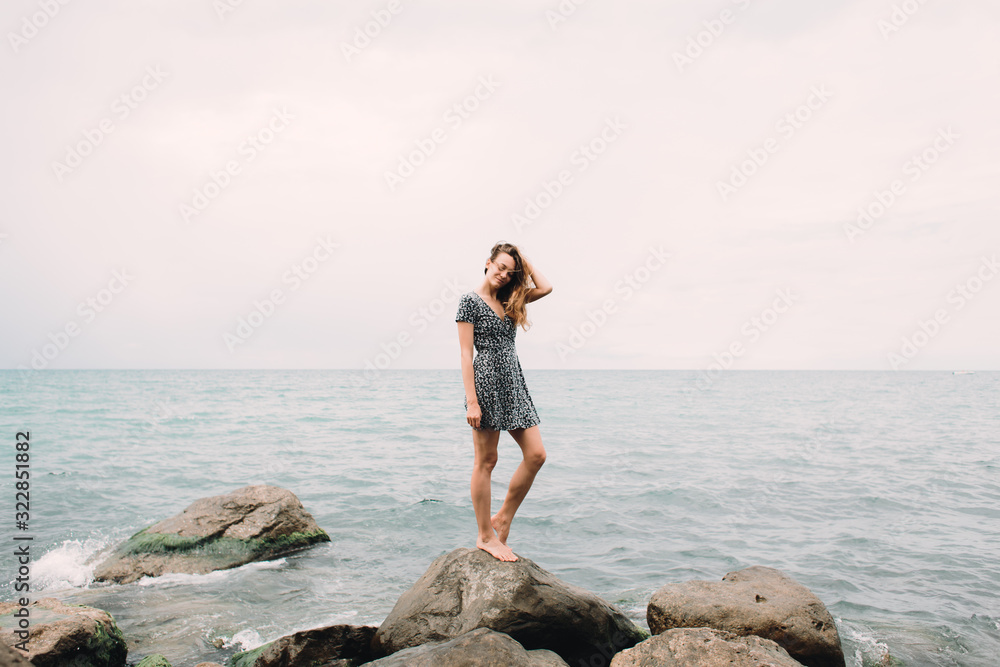 a young girl in a dress stands on the beach on large stones, the wind develops her hair