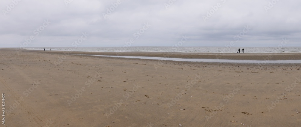 Sandy beach on the North Sea, almost deserted, only a few lonely walkers, traces in the sand, only small waves on the gray water Sandy beach on the North Sea, almost deserted, only a few lonely walkers, traces in the sand, only small waves on the gray water
